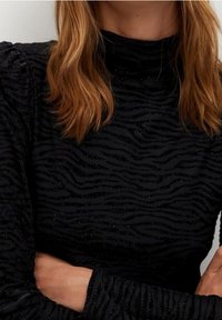 Femme aux cheveux blonds mi-longs portant un haut noir à manches longues avec une subtile texture à rayures zèbre, les bras croisés à la taille.