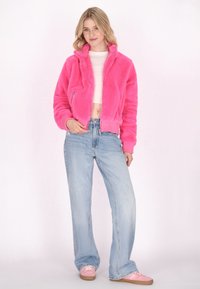 Giacca bomber rosa fluffy con zip, polsini a coste e design cropped, abbinata a jeans a gamba larga blu chiaro e sneakers rosa.