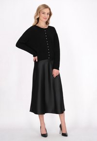 Černý cardigan s detailním zapínáním, zkombinovaný s dlouhou černou saténovou sukní a černými podpatky. Hladké textury a klasický silueta.