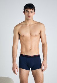 Marineblaue Boxer Shorts mit rotem "HUGO"-Logo auf einem schwarzen Bund, aus einem weichen, dehnbaren Material, enganliegendes Design, mit glatter Textur.