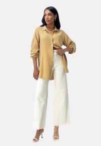 Beige strukturierte Hemdbluse mit aufgerollten Ärmeln, kombiniert mit hochtaillierten weißen weit geschnittenen Hosen und beigen Riemchen-Heels.