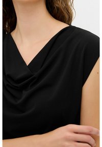 Mujer vestida con un top negro sin mangas con un escote drapeado, que muestra parte de su cuello, hombro y mano.