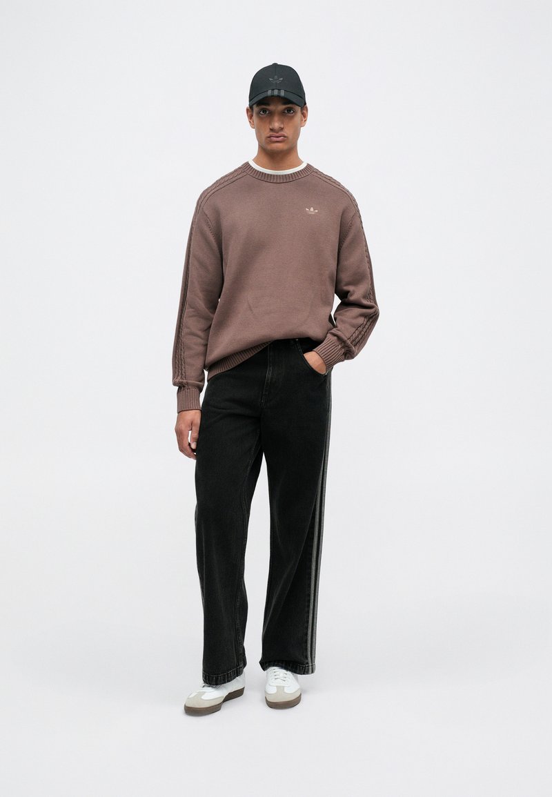 Brown knitted sweater with ribbed edges, black high-waisted jeans with side stripes, and white sneakers. Black cap with logo completes the outfit.