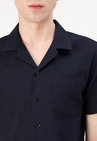 Camicia a maniche corte blu navy con trama testurizzata, colletto a punte e taschino sul petto. Cinque bottoni neri davanti.