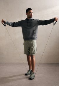 Grå hættetrøje, olivengrønne shorts og grønne træningssko. Holder modstandsbånd, arme strakt ud, mod en neutral baggrund.