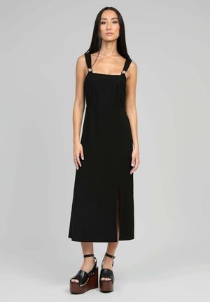 Robe noire midi avec encolure carrée, larges bretelles avec des accents métalliques, et une fente sur le côté. Associée à des sandales plateformes noires chunky.
