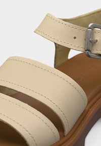 Μπεζ δερμάτινο σανδάλι με δύο φαρδιές λουρίδες κατά μήκος του ποδιού, λουρίδα στον αστράγαλο με ασημένια αγκράφα και καφέ μαλακή σόλα.