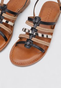 Sandalias de cuero marrón con tiras tejidas negras y con patrón, suela plana con detalles de costura y diseño de punta abierta.