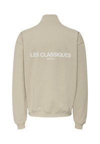 Jasno beżowy sweatshirt z wysokim kołnierzem, z białym napisem "LES CLASSIQUES" i "ORIGINAUX" na plecach. Miękka tkanina, ściągacze na rękawach.