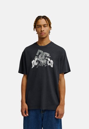 Młody mężczyzna z warkoczykami stoi przodem, ubrany w luźny czarny t-shirt DC SHOES z nadrukiem i niebieskie dżinsy, na jednolitym białym tle.