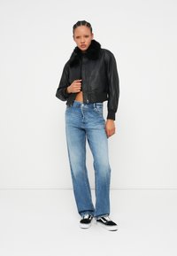 Kobieta z zaplecionymi włosami, ubrana w czarną skórzaną kurtkę z futrzanym kołnierzem, jasnoniebieskie dżinsy i czarno-białe sneakersy, stojąca na białym tle.