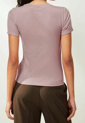 Frau trägt ein kurzärmeliges, mauvefarbenes, geripptes Strickoberteil und dunkelbraune Hose, von hinten vor einem schlichten hellen Hintergrund gezeigt.