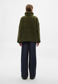 Õliva rohelise fliisjaki kõrge krae, lõdva lõike ja tekstuurse pinnaga, kombineeritud tumedate denimist laia säärega pükstega.