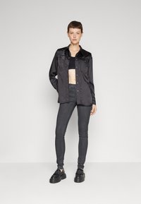 Chaqueta de camisa de satén negro con cuello puntiagudo, cierre de botones, dos bolsillos en el pecho, combinada con pantalones ajustados negros y zapatos negros gruesos.