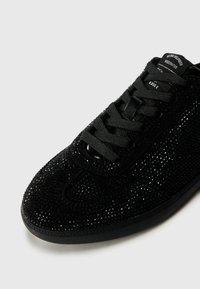 Baskets noires avec une surface texturée ornée de nombreux petits strass, à lacets sur le devant et à bout rond. Semelle en caoutchouc et minimalisme au niveau du logo.