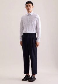 Camicia bianca con piccoli pois blu navy, collo con bottoni e maniche lunghe; abbinata a pantaloni plissettati blu navy e scarpe nere con lacci.