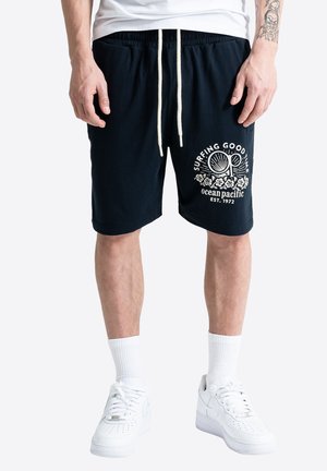 Mężczyzna noszący czarne szorty z białym logo "Surfing Good Time Ocean Pacific", białe skarpetki i białe trampki, stojący na jednolitym tle.
