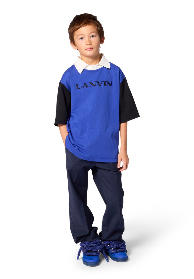 Αγόρι που φοράει μπλε και μαύρο πουκάμισο Lanvin, σκούρο παντελόνι και μπλε αθλητικά με υπερμεγέθεις χοντρές μπλε κορδόνια, στέκεται μπροστά σε λευκό φόντο.