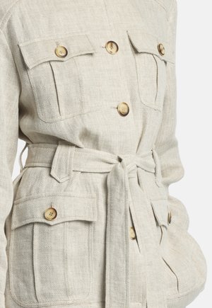 Béžový sako so vzorom rybej kosti, so štyrmi zapínacími prednými vreckami s chlopňami a zodpovedajúcim látkovým opaskom previazaným v páse.