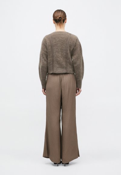 Femeie cu părul prins în coc, purtând un pulover maro pufoasă și pantaloni maro cu talie înaltă și picioare largi, stând cu fața spre un fundal alb.