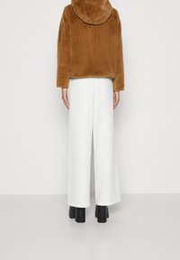 Chaqueta marrón de piel sintética con capucha, combinada con pantalones blancos de pierna ancha y botines negros de plataforma, vista desde atrás.