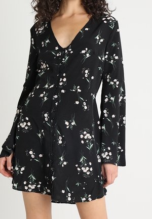 Kobieta ubrana w czarną, kwiatową sukienkę na guziki z długimi rękawami i głębokim dekoltem w kształcie litery V na jednolitym tle.