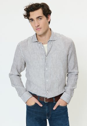 Jeune homme aux cheveux bouclés bruns portant une chemise rayée grise et blanche, un jean bleu et une ceinture marron, les mains dans les poches, sur fond blanc.