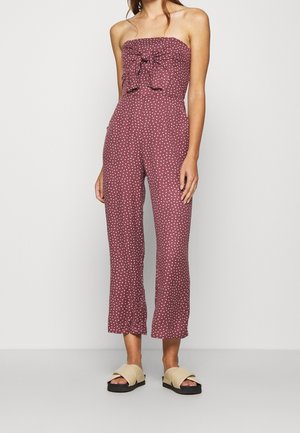 Kobieta ubrana w bezramkowy kombinezon w kolorze mauve z białymi kropkami, z przodu kokarda, szerokie, skrócone nogawki i beżowe sandały wsuwane.