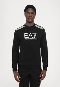 Black sweatshirt with long sleeves, ribbed collar, and cuffs. Features a white EA7 Emporio Armani logo on the front and stripe accents on the shoulders.