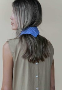 Haut sans manches beige avec une fermeture à boutons au dos, associé à une écharpe rayée bleue nouée au cou. Texture douce et lignes épurées.