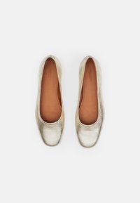 Metalliske gullflats med rund tå, glatt tekstur og en tanfarget innvendig fôr. Enkel design uten synlig maskinvare eller accenter.
