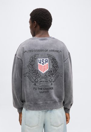 Personne portant un sweat-shirt gris délavé avec un écusson des États-Unis, une couronne de laurier et le texte "United States of America" et "Be the Change USMNT" dans le dos.