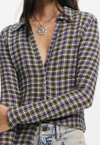 Chemise à carreaux avec un mélange de motifs verts, violets et blancs. Dispose d'un devant boutonné, d'un col et d'un tissu texturé. Portée avec un jean en denim bleu.