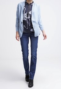 Světlá modrá džínová bunda přes tmavě modrou košili s bílými vzory plameňáků, doplněná tmavě modrými džínami s rovnými nohavicemi a černými kotníkovými botami.