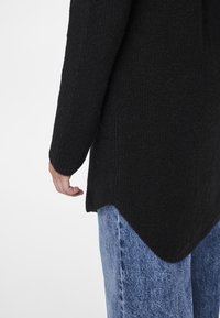 Schwarzer Strickpullover mit strukturierter Oberfläche und geschwungenem Saum. Kombiniert mit locker sitzenden hellblauen Jeans aus Denim.