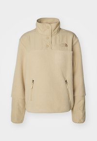 Beżowy polarowy sweter z wysokim kołnierzem, zapięciem na napy, dwoma kieszeniami na zamek i pikowanym detalem na ramionach. Miękka tekstura w całej powierzchni.