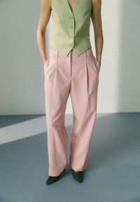 Osoba w jasnozielonej kamizelce z guzikami, luźnych różowych spodniach o wysokim stanem i spiczastych oliwkowych butach, stoi z rękami w kieszeniach.