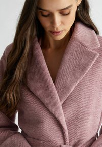 Cappotto di lana rosa pallido con motivo a spina di pesce, ampie revers e vita accentuata da una cintura, caratterizzato da una finitura testurizzata e un tessuto morbido.