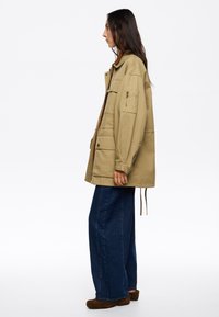 Chaqueta caqui con grandes bolsillos, cierre de botones y dobladillo ajustable, combinada con unos pantalones de mezclilla azules holgados y zapatos marrones.