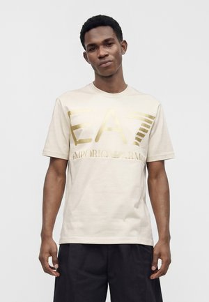 Jeune homme portant un t-shirt beige clair avec un grand logo doré EA7 Emporio Armani, associé à un pantalon noir, debout devant un fond uni.