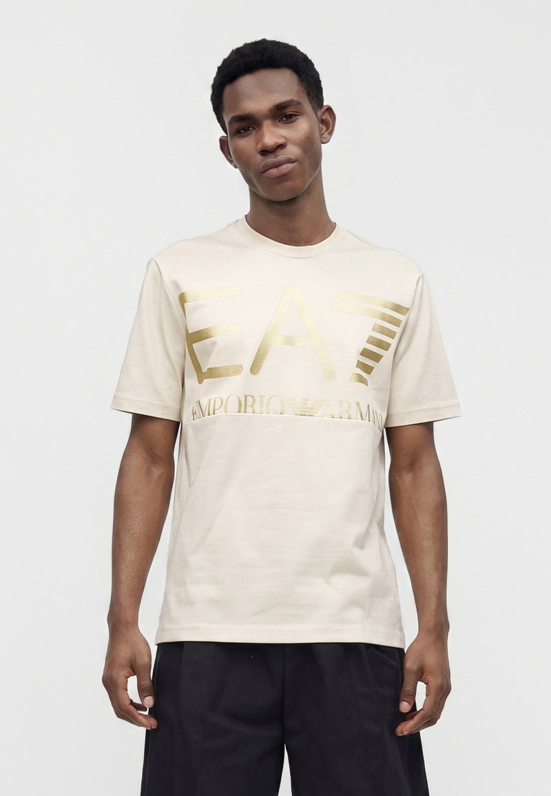 Νεαρός άνδρας που φορά ανοιχτό μπεζ T-shirt με μεγάλο χρυσό λογότυπο EA7 Emporio Armani, συνδυασμένο με μαύρο παντελόνι, στέκεται μπροστά από απλό φόντο.
