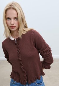 Cardigan lavorato a maglia marrone con orlo arricciato, chiusure a bottone e maniche lunghe. La texture è a coste e il design complessivo è aderente.