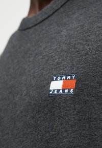 Γκρι βαμβακερό t-shirt με ορθογώνιο κεντητό λογότυπο σε κόκκινο, άσπρο και μπλε με το κείμενο "TOMMY JEANS" στο αριστερό μέρος του στήθους.