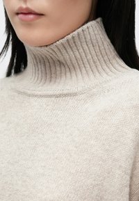Gros plan d'une personne portant un pull à col roulé côtelé beige avec de longs cheveux foncés partiellement visibles.