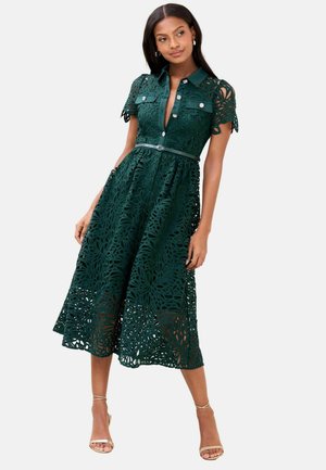 Dunkelgrünes Spitzenkleid mit Hemdkragen, kurzen Ärmeln, floralem Ausschnittmuster, durchgehender Knopfleiste und passendem Gürtel in der Taille.