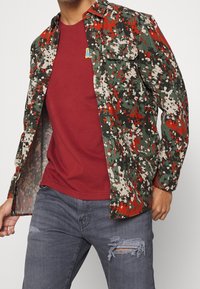 Overhemd met knopen in een groen, rood en beige camouflagepatroon, met lange mouwen en twee borstzakken. Draagt over een wijnrode shirt.