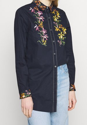 Chemise bleu marine à boutons avec broderies florales roses, jaunes et orange sur le devant, le col et les poignets, portée avec un jean bleu clair.