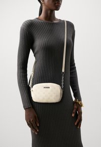 Hvid læder crossbody taske med præget logo, rund form, justerbar strop og kædedetalje. Båret over en ribbet grå kjole.