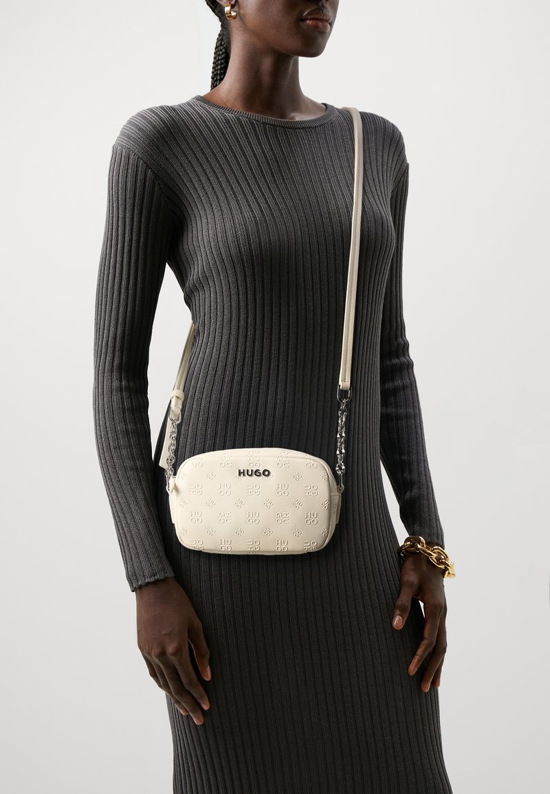 Hvid læder crossbody taske med præget logo, rund form, justerbar strop og kædedetalje. Båret over en ribbet grå kjole.