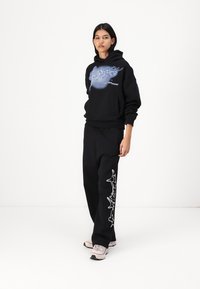 Sweatshirt preta com um design gráfico azul na parte da frente. Calças de moletom pretas com padrões abstratos brancos. Ténis de tom claro.